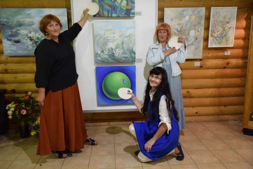 VIZĪTKARTE. Visas trīs mākslinieces vienādā formātā izveidojušas katra savas zīmes raksturojumu gleznā. Gunta Vēvere (no kreisās) ir pastiepusi roku pret uguns zīmi, Mendija Leoke (vidū) ir ūdens zīme, Inese Dembovska — zemes zīme. Visām gleznotājām rokā ir Mendijas gatavotās paletes.